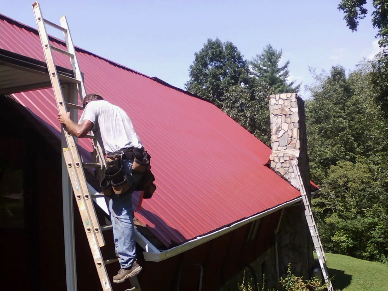Roofer on a ladder beside a red metal roof and stone chimney — Soffit & Fascia Repair by Roofing Brothers in Wakita
